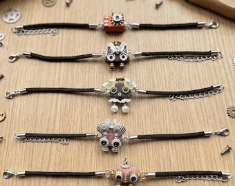 Könnte beinhalten: Fünf Armbänder mit schwarzen Kordelbändern und silberfarbenen Metallverschlüssen. Jedes Armband hat einen einzigartigen zentralen Anhänger in Form eines kleinen Roboters oder Wesens, verziert mit verschiedenen Elementen wie Zahnrädern, buntem Emaille und kleinen weißen Perlen.