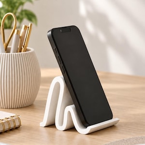 May include: A white smartphone stand with a wavy, serpentine silhouette holding a black smartphone in a vertical position. The stand features a smooth, matte finish and a curved design that supports the device at an angled viewing position.