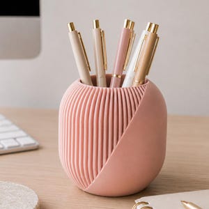 May include: A cylindrical pink pen holder with a ribbed texture on one side and a smooth, diagonal section on the other. The holder contains several pens with gold-colored clips and caps, including shades of beige, white, and muted pink.