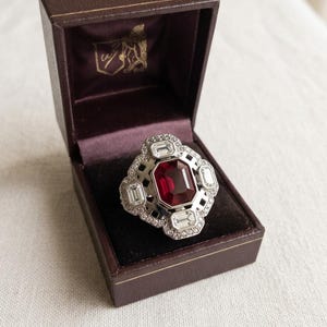 Peut inclure: Une bague argentée ornée d'une grande pierre rectangulaire rouge foncé, entourée de quatre pierres rectangulaires transparentes plus petites et d'un bord de minuscules pierres rondes transparentes, présentée dans un écrin marron foncé.