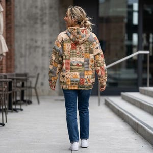 Puede incluir: Una sudadera con capucha y manga larga que presenta un diseño de patchwork con varios cuadrados estampados que incluyen motivos florales, graneros rojos, girasoles y texturas de cuadros en tonos verde, amarillo, rojo y crema, combinada con vaqueros azules.