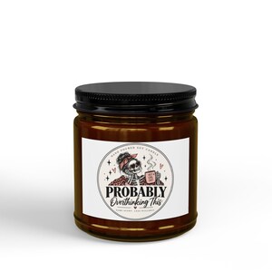 May include: A brown glass candle jar with a black lid and a white circular label. The label features a skeleton illustration with a pink headband, holding a mug labelled Hot Girl Shit, with the text Probably Overthinking This and Hand Poured Soy Candle.