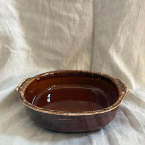 Pode incluir: Uma assadeira redonda de cerâmica castanho-escura com duas pequenas pegas nos lados opostos. O interior e o exterior apresentam um vidrado brilhante de cor castanha profunda, enquanto o rebordo tem um padrão decorativo texturado em creme.