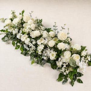 Könnte beinhalten: Ein langes Blumenarrangement mit weißen Rosen, Hortensien und kleinen Füllblumen, durchsetzt mit grünen Eukalyptusblättern und verschiedenem Blattwerk. Die Komposition ist dicht und länglich und zeigt eine Mischung aus weißen Blüten.