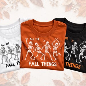 Puede incluir: Tres camisetas de cuello redondo en blanco, naranja quemado y negro. Cada camiseta presenta un estampado gráfico de cuatro esqueletos bailando y tocando instrumentos musicales con el texto "ALL THE FALL THINGS" en una fuente negrita blanca o negra.