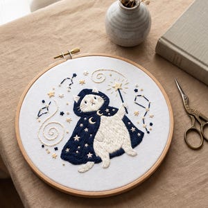 Puede incluir: Un bastidor de bordado circular de madera que contiene tela blanca con un diseño cosido a mano de un gato blanco con una capa azul oscuro con capucha. El gato sostiene una varita con punta de estrella, rodeado de constelaciones, estrellas y espirales.