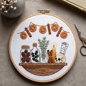 Puede incluir: Un bastidor de bordado circular con un panel de tela blanca y un diseño cosido a mano. El bordado muestra un estante de madera con frascos de piñas, acebo, romero, ramas de canela, una vela encendida y una calavera con un mapa estelar.