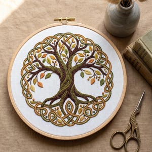 May include: A circular wooden embroidery hoop containing white fabric with a hand-stitched tree design. The tree features a dark brown trunk and branches, surrounded by a braided border in shades of green, gold, and brown, with small leaves.