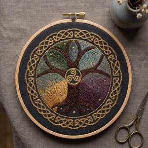 May include: A circular wooden embroidery hoop containing a dark fabric panel with a hand-stitched tree design. The tree features four distinct colored quadrants of foliage, a central gold triskelion symbol, and a gold embroidered Celtic knot border.