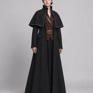 May include: A floor-length black coat featuring a high collar, long sleeves, and a detachable capelet with white contrast stitching. Underneath, a brown suede vest is worn with a brown leather belt featuring a gold-toned buckle and a cross-body strap.