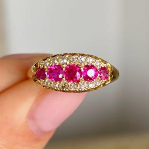 Puede incluir: Un anillo de oro con una fila horizontal de cinco piedras preciosas redondas de color rosa vibrante en el centro. Las piedras están rodeadas por un borde de pequeñas piedras blancas claras y brillantes incrustadas en un engaste de oro ovalado.