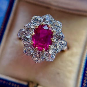 May include: A cluster ring featuring a central oval-cut vibrant red gemstone surrounded by a halo of ten round-cut clear white diamonds. The stones are set in a metal band, creating a floral-inspired arrangement with a high-contrast color palette.