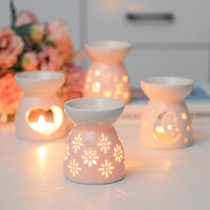 May include: Three light pink ceramic oil burners with cut-out designs. One features a heart shape, one a moon and stars pattern, and the front one displays a floral motif. Each burner has a bowl at the top and a hollow base for a tea light candle.