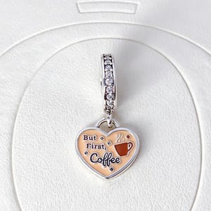 Könnte beinhalten: Ein herzförmiger silberner Anhänger mit hellpfirsichfarbenem Emaille-Hintergrund, der den Schriftzug "But First, Coffee" in schwarzer Schreibschrift, ein kleines braunes Kaffeetassen-Symbol und klare Zirkonia-Steine am oberen Bügel aufweist.