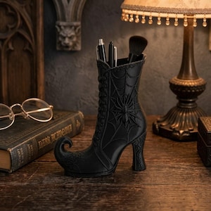 May include: A black desk organizer shaped like a Victorian-style lace-up boot with a curled toe. The boot features a raised spider and web design on the side, a high heel, and holds two pens and a makeup brush in its open top opening.