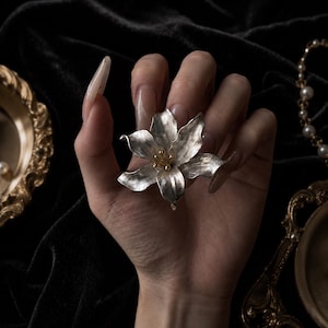 May include: A silver-toned flower brooch with textured petals and a central cluster of small gold-toned stamens. The brooch features a realistic floral design with six distinct petals radiating from the center, showcasing a metallic finish.