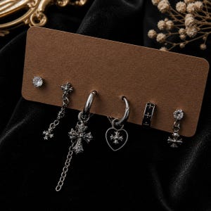 May include: A set of five silver-toned earrings displayed on a brown card. The collection includes two crystal studs, one cross-dangle earring with a chain, one hoop earring with a heart-shaped cross charm, and one small band earring with cross motifs.