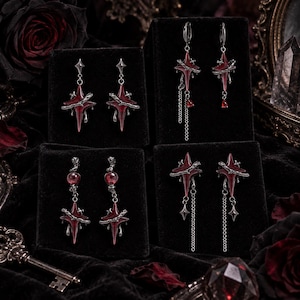 Könnte beinhalten: Vier Paar Ohrringe mit sternförmigen Anhängern aus dunklem Metall, tiefroten Emaille-Zentren und silberfarbenen dornartigen Verzierungen. Einige Designs enthalten feine silberne Ketten, kleine rote tropfenförmige Kristalle oder runde rote Perlen.