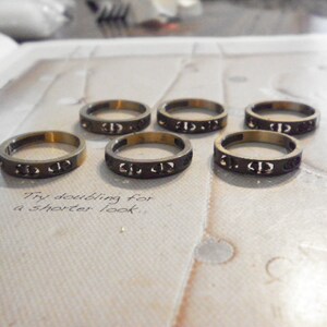 Puede incluir: Seis anillos de color bronce con un diseño recortado repetido se muestran sobre una superficie clara. Los anillos son circulares y metálicos. El texto "Try doubling for a shorter look" está escrito debajo. Joyas.