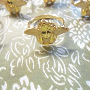 May include: Gold-coloured rings with a distinctive design featuring a face with outstretched wings. The rings are arranged on a patterned green and white surface, creating a visually appealing display. The rings have a shiny finish.