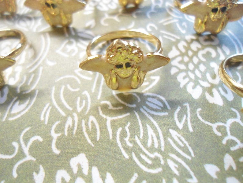 May include: Gold-coloured rings with a distinctive design featuring a face with outstretched wings. The rings are arranged on a patterned green and white surface, creating a visually appealing display. The rings have a shiny finish.