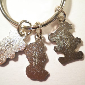 K&ouml;nnte beinhalten: Ein silberfarbener Schl&uuml;sselanh&auml;nger mit drei kleinen, b&auml;renf&ouml;rmigen Anh&auml;ngern. Die Anh&auml;nger sind an einem gro&szlig;en Ring befestigt. Die Anh&auml;nger haben eine strukturierte Oberfl&auml;che und sind mit kleinen Details versehen.