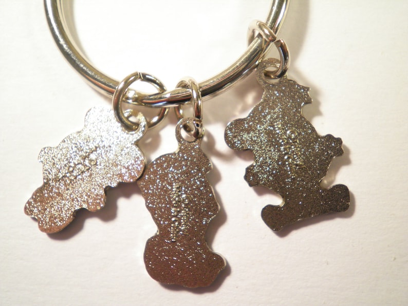 K&ouml;nnte beinhalten: Ein silberfarbener Schl&uuml;sselanh&auml;nger mit drei kleinen, b&auml;renf&ouml;rmigen Anh&auml;ngern. Die Anh&auml;nger sind an einem gro&szlig;en Ring befestigt. Die Anh&auml;nger haben eine strukturierte Oberfl&auml;che und sind mit kleinen Details versehen.