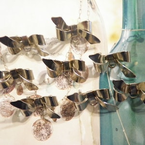 May include: Several silver-toned bow necklaces with delicate chains and round, textured pendants. The necklaces are displayed in front of a blurred background featuring glass bottles. The bows have a shiny, metallic finish.