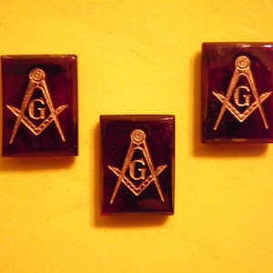 Puede incluir: Tres pines rectangulares con emblemas masónicos. Cada pin presenta un símbolo de escuadra y compás de color plateado con la letra "G" en el centro, sobre un fondo rojo oscuro. Los pines se muestran sobre una superficie amarilla.