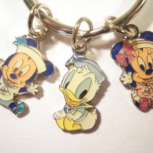 K&ouml;nnte beinhalten: Ein silberfarbenes Armband mit drei Emaille-Anh&auml;ngern mit den Zeichentrickfiguren Mickey Mouse, Donald Duck und Minnie Mouse. Die Anh&auml;nger sind im Seemanns-Thema gehalten, mit blauen, wei&szlig;en, gelben und rosa Akzenten.