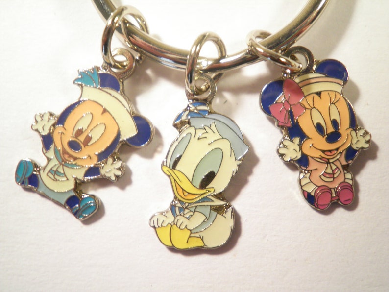 K&ouml;nnte beinhalten: Ein silberfarbenes Armband mit drei Emaille-Anh&auml;ngern mit den Zeichentrickfiguren Mickey Mouse, Donald Duck und Minnie Mouse. Die Anh&auml;nger sind im Seemanns-Thema gehalten, mit blauen, wei&szlig;en, gelben und rosa Akzenten.