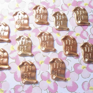May include: A collection of bronze-colored metal charms, each shaped like a scroll with the letters "INRI" inscribed. The charms have a small hole at the top and are arranged on a floral patterned background.