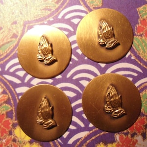 Könnte beinhalten: Vier goldfarbene, runde Metallknöpfe mit einem erhabenen Design von betenden Händen. Die Knöpfe sind auf einem gemusterten Hintergrund aus Lila, Weiß und floralen Designs angeordnet.