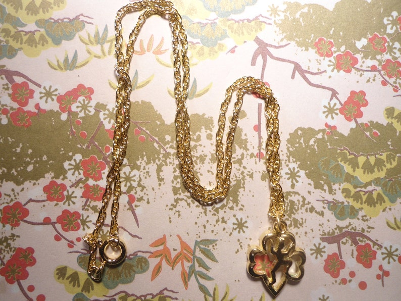 May include: Gold-coloured necklace with a pendant featuring a floral design. The chain is a twisted rope style. The pendant has a gold outline with a pink and orange floral design inside. The necklace is displayed on a floral patterned background.
