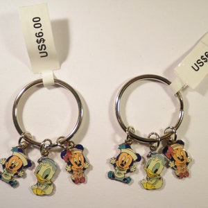 K&ouml;nnte beinhalten: Zwei Schl&uuml;sselanh&auml;nger mit silbernen Ringen, jeweils mit drei Cartoon-Charms: Mickey Mouse, Donald Duck und Minnie Mouse. Die Charms sind farbenfroh, mit blauen, gelben, roten und wei&szlig;en Akzenten. Jeder Schl&uuml;sselanh&auml;nger hat ein Etikett mit der Aufschrift "US$6.00".