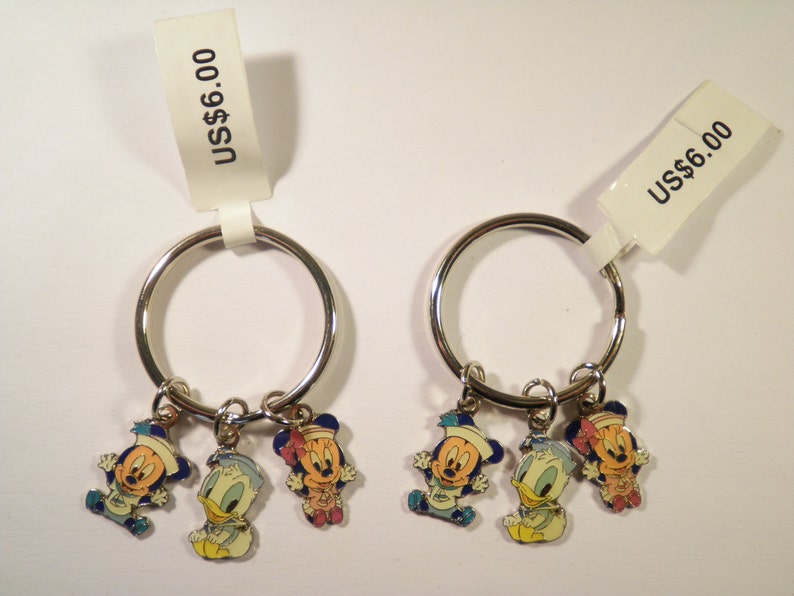 K&ouml;nnte beinhalten: Zwei Schl&uuml;sselanh&auml;nger mit silbernen Ringen, jeweils mit drei Cartoon-Charms: Mickey Mouse, Donald Duck und Minnie Mouse. Die Charms sind farbenfroh, mit blauen, gelben, roten und wei&szlig;en Akzenten. Jeder Schl&uuml;sselanh&auml;nger hat ein Etikett mit der Aufschrift "US$6.00".