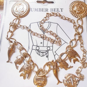 May include: Gold-toned metal waist belt with a chain design and decorative charms. The belt features round medallions and various sea creature charms, including dolphins and fish. The belt is displayed on a white card with a line drawing of a dress.