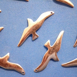 May include: A collection of silver-coloured metal bird charms are arranged on a blue surface. Each charm is shaped like a stylized bird in flight, with a small hole for threading. The charms have a smooth, polished finish.