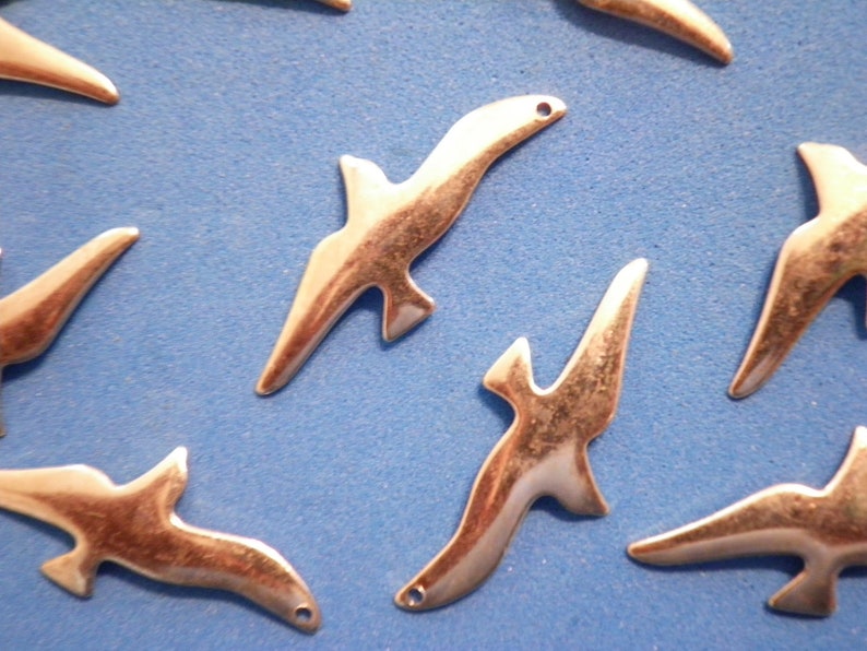 May include: A collection of silver-coloured metal bird charms are arranged on a blue surface. Each charm is shaped like a stylized bird in flight, with a small hole for threading. The charms have a smooth, polished finish.