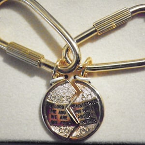 Puede incluir: Llavero de metal dorado con un colgante dividido. El colgante presenta una inscripción religiosa. El llavero tiene un cierre de mosquetón y un cierre de rosca. El colgante está en dos mitades, unidas por un pequeño anillo de metal.