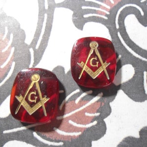 May include: Two square, red-toned Masonic cufflinks with gold-colored symbols. The symbols include a compass, a square, and the letter "G". The cufflinks have a marbled appearance and are set against a patterned background.