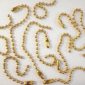 May include: Several strands of gold-colored ball chain with matching connectors are arranged on a white surface. The chains are made of small, round beads linked together, with elongated connectors at the ends.