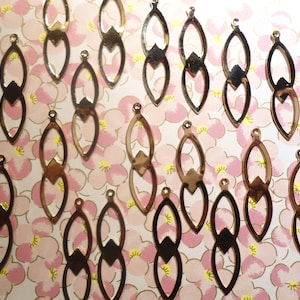 May include: An assortment of bronze-colored earring charms. Each charm features an elongated oval shape connected to a diamond-shaped accent. The charms are arranged on a floral patterned background.