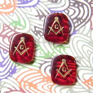 May include: Three square-shaped, red-toned buttons with a gold-colored Masonic symbol. The symbol includes a compass, a square, and the letter "G". The buttons have a glossy finish and are set against a colorful, patterned background.