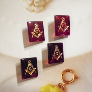 May include: Four square, burgundy-colored Masonic emblem charms. Each charm features a gold-colored symbol of a compass and square with a letter "G" in the center. The charms are set against a white background.