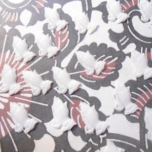 May include: White praying hands charms scattered on a patterned background. The charms are small, with detailed fingers and a smooth, glossy finish. The background features a floral design in shades of gray, white, and burgundy.