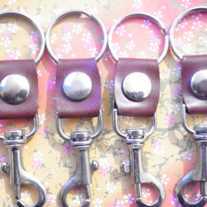 May include: Four keychains with burgundy leather straps and silver-colored metal hardware. Each keychain has a large metal ring, a snap hook, and a circular metal button. The keychains are arranged in a row.