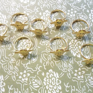 May include: A selection of gold-coloured metal rings, each with a decorative feature. The rings are displayed on a patterned surface. The decorative element appears to be a stylised face with outstretched wings.