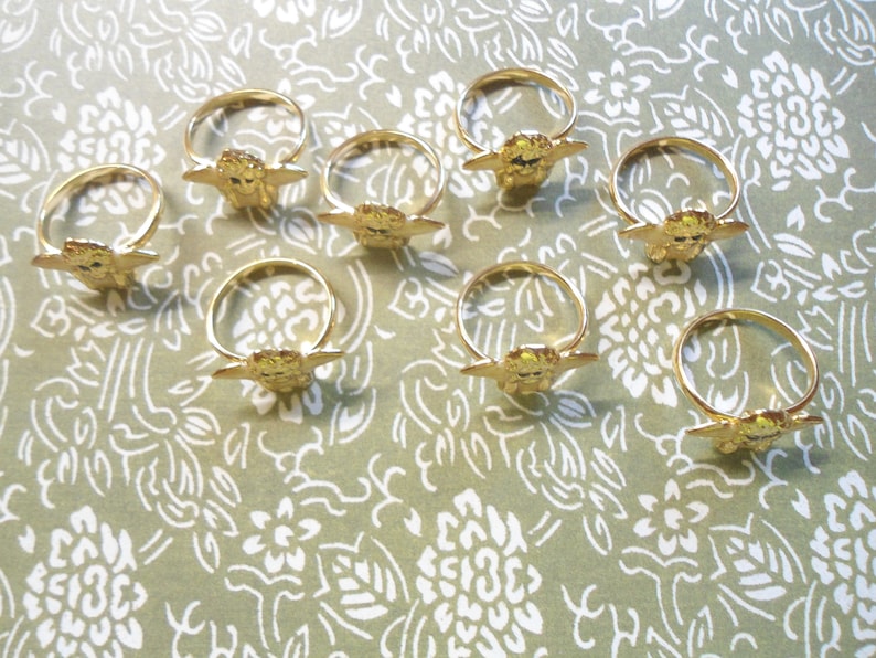 May include: A selection of gold-coloured metal rings, each with a decorative feature. The rings are displayed on a patterned surface. The decorative element appears to be a stylised face with outstretched wings.