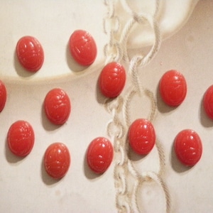 May include: A collection of eleven oval, red scarab beads. Each bead has a carved design and a glossy finish. The beads are arranged on a light-colored surface with a subtle pattern, suggesting a vintage or antique aesthetic.